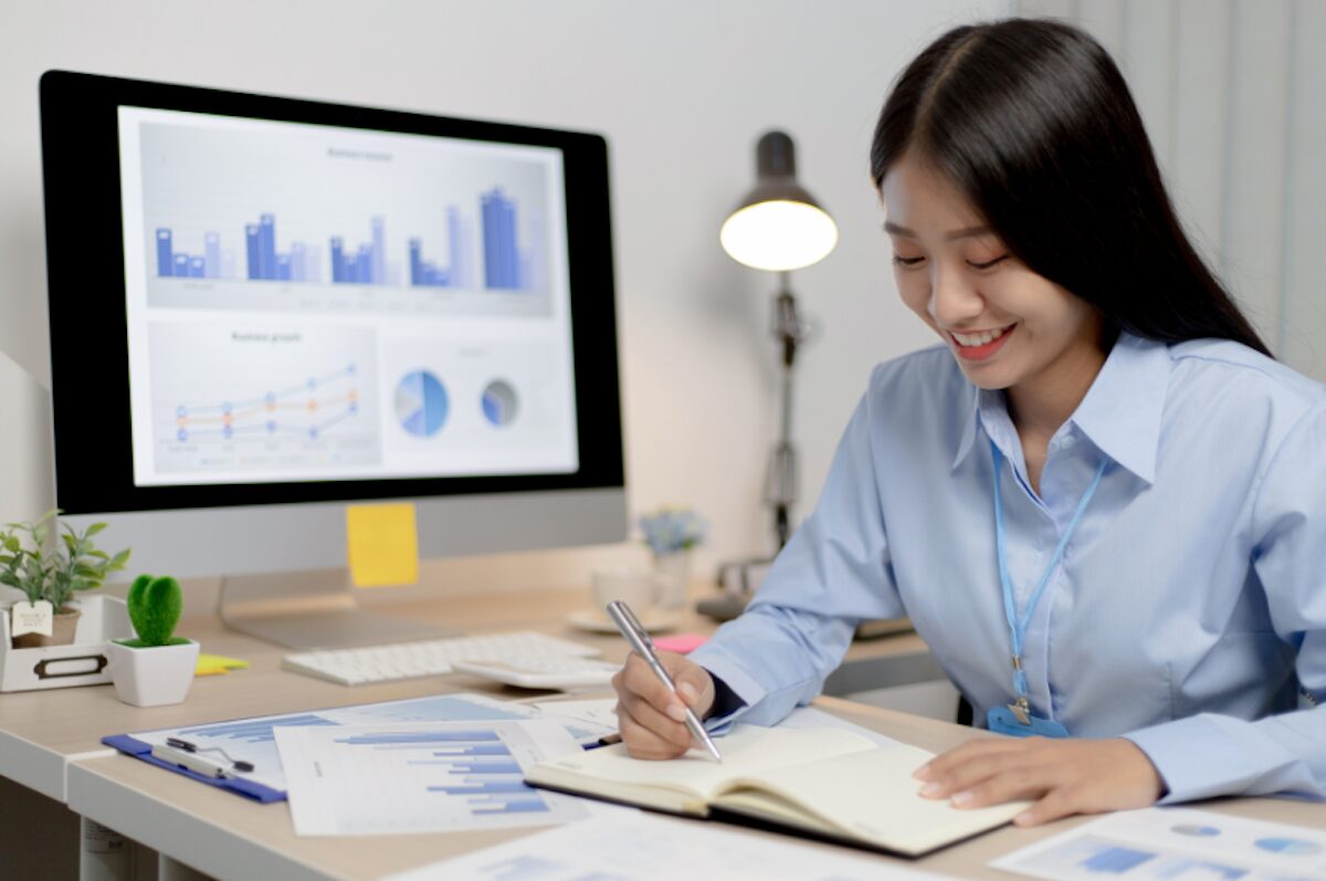 A businesswoman reviewing charts and graphs on a monitor while taking notes